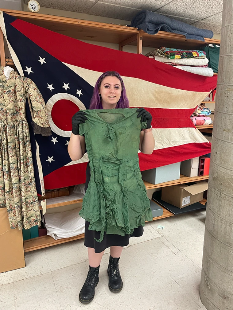 Milana holds a dress she is cataloguing during her training at Arizona History Museum. She stands in front of a historical flag.